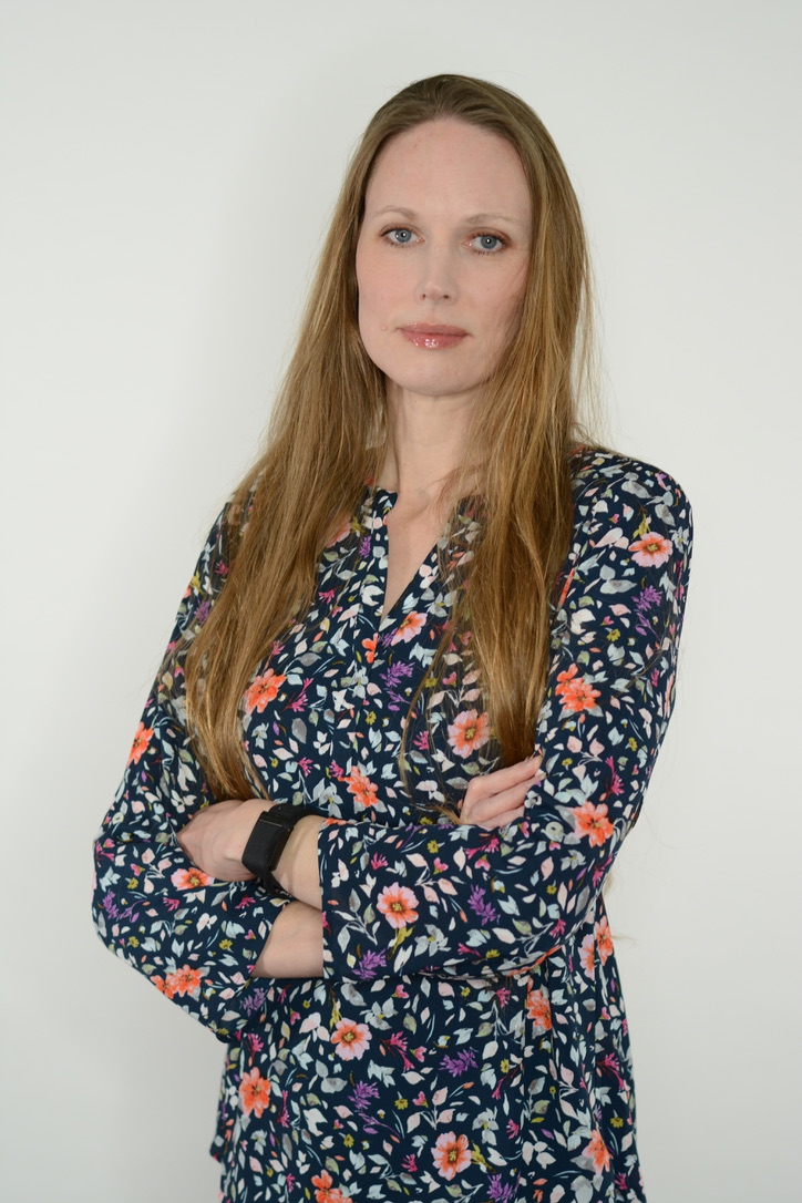 Cassandra a white woman with long dark blonde hair wears a floral pattern shirt and stands in front of a cream colored wall.